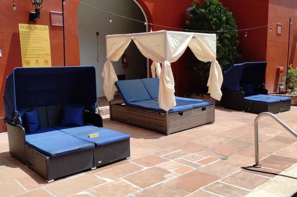 Outdoor poolside lounging area with canopy beds, blue cushions, and red brick wall backdrop