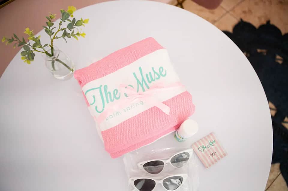 White table with pink towel branded 'The Muse', sunglasses, and small toiletries on display
