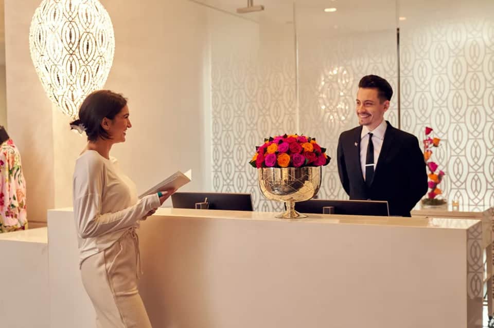 Hotel front desk with receptionist assisting guest, featuring white counter and vibrant floral arrangement