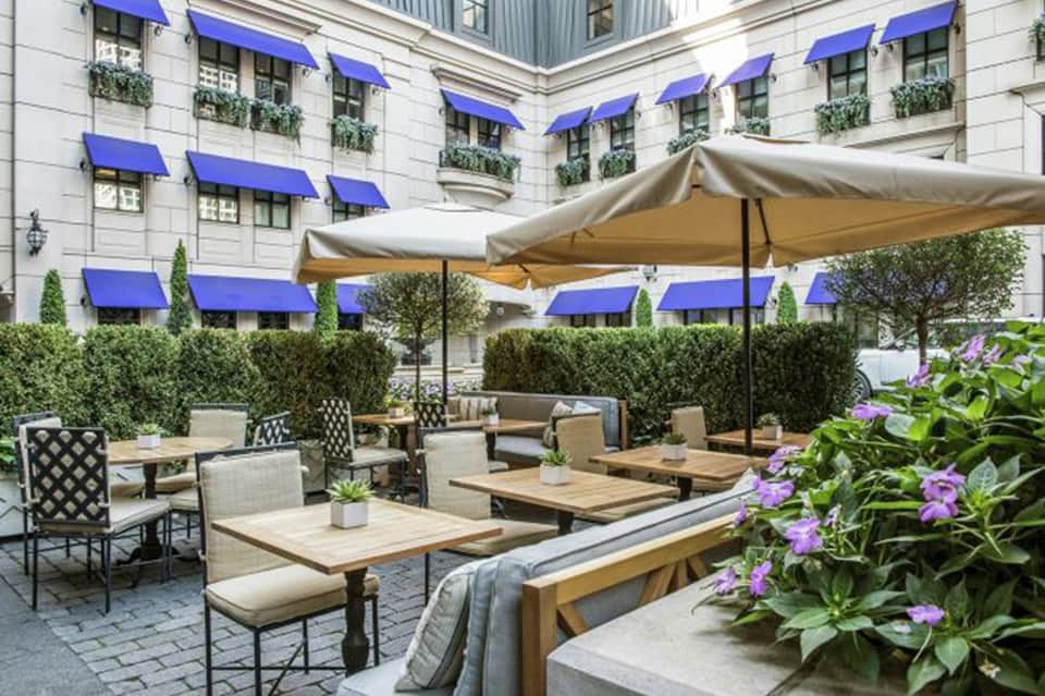 Elegant courtyard with blue umbrellas, wooden dining tables, lounge seating, and manicured hedges