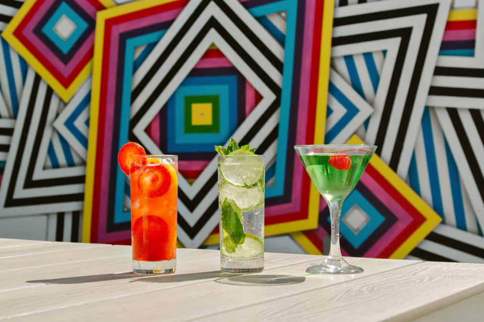 Three colorful cocktails displayed against vibrant geometric background mural