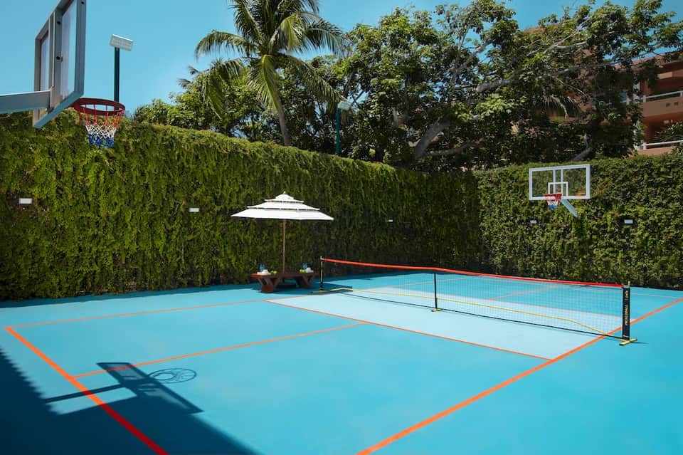 Blue multi-sport court with tennis net and basketball hoop, surrounded by green hedges and white umbrellas