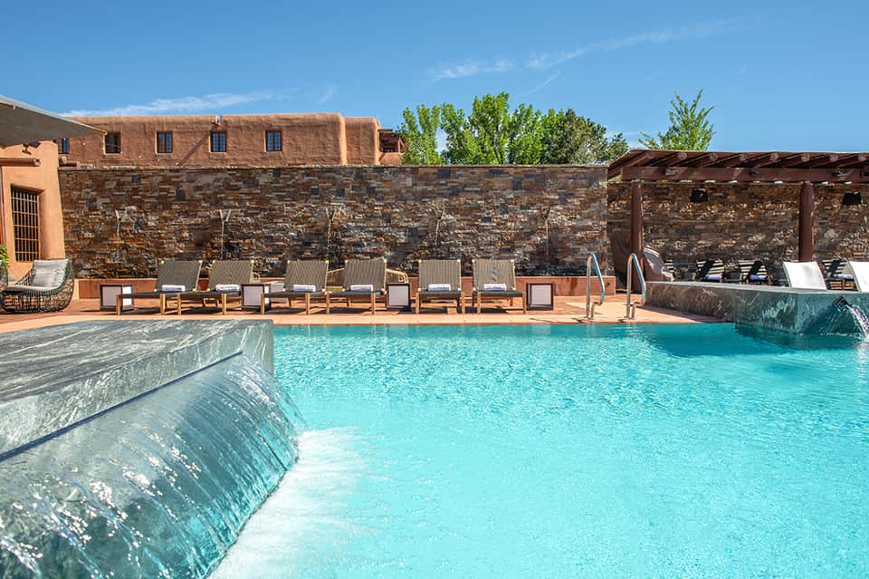Resort pool with turquoise water, stone walls, lounge chairs, pergola shade structures, and desert landscape