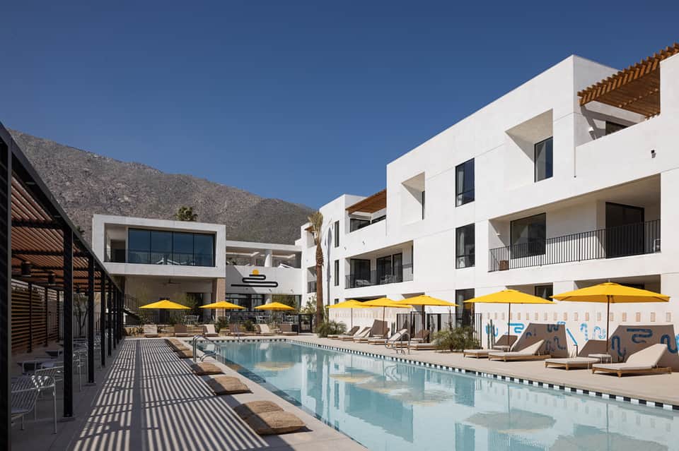 Modern resort pool with white building architecture, yellow umbrellas, loungers, and mountain backdrop under clear sky