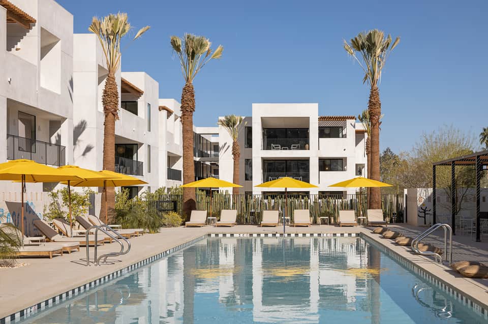 Modern white apartment complex with pool and yellow umbrellas, featuring contemporary architecture and palm trees