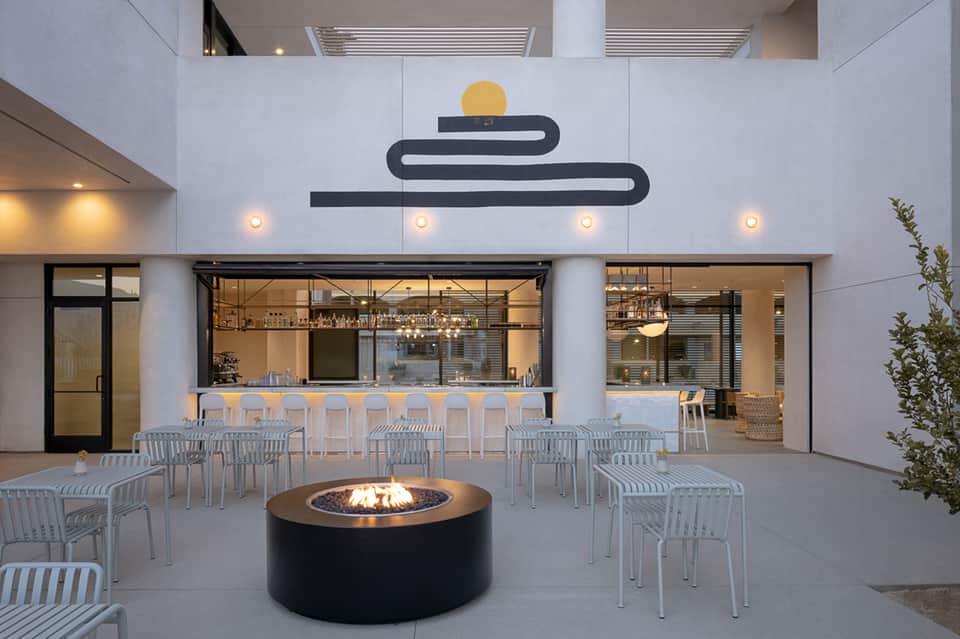 Modern courtyard with minimalist bar, fire pit, white chairs, and abstract black wall art with yellow accent