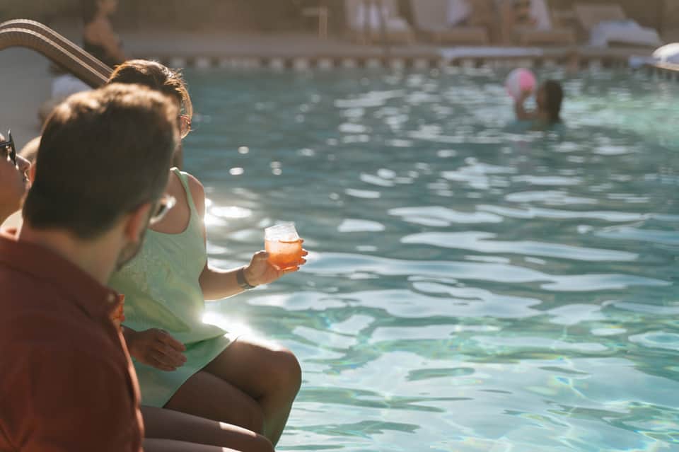 Woman sitting poolside holding an orange cocktail while watching swimmers