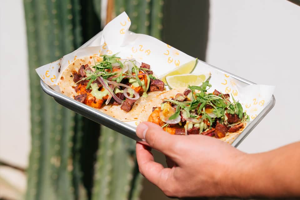 Hand holding plate of two tacos with meat, fresh vegetables, lime wedges, and cilantro garnish