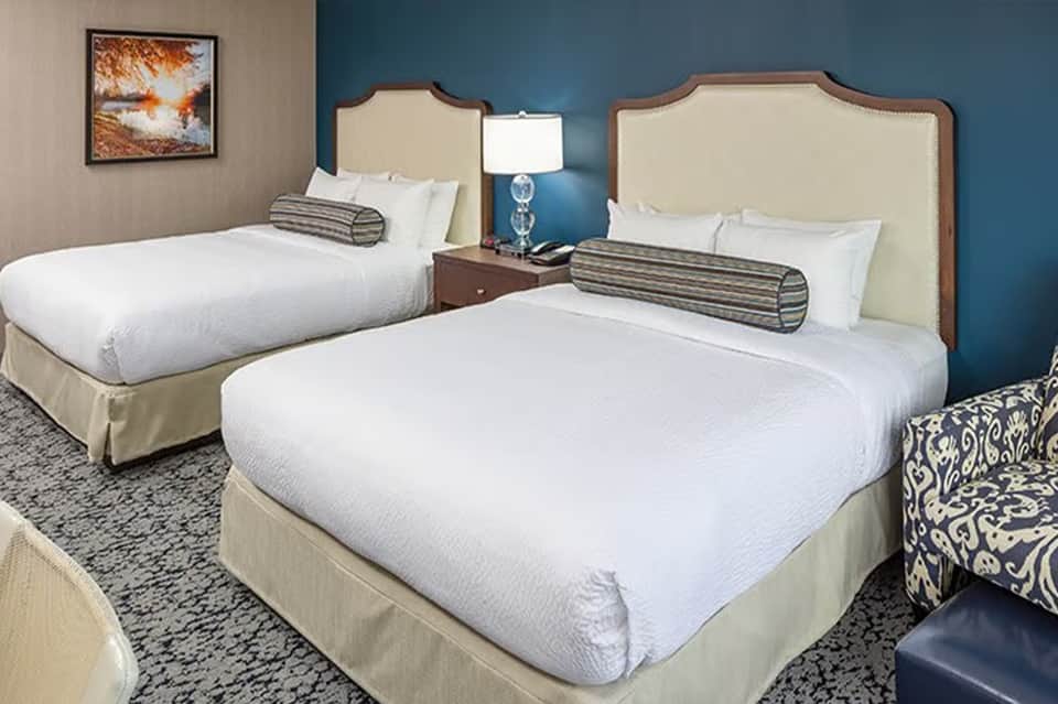 Hotel room with two queen beds, cream upholstered headboards, blue accent wall, and bedside lamp