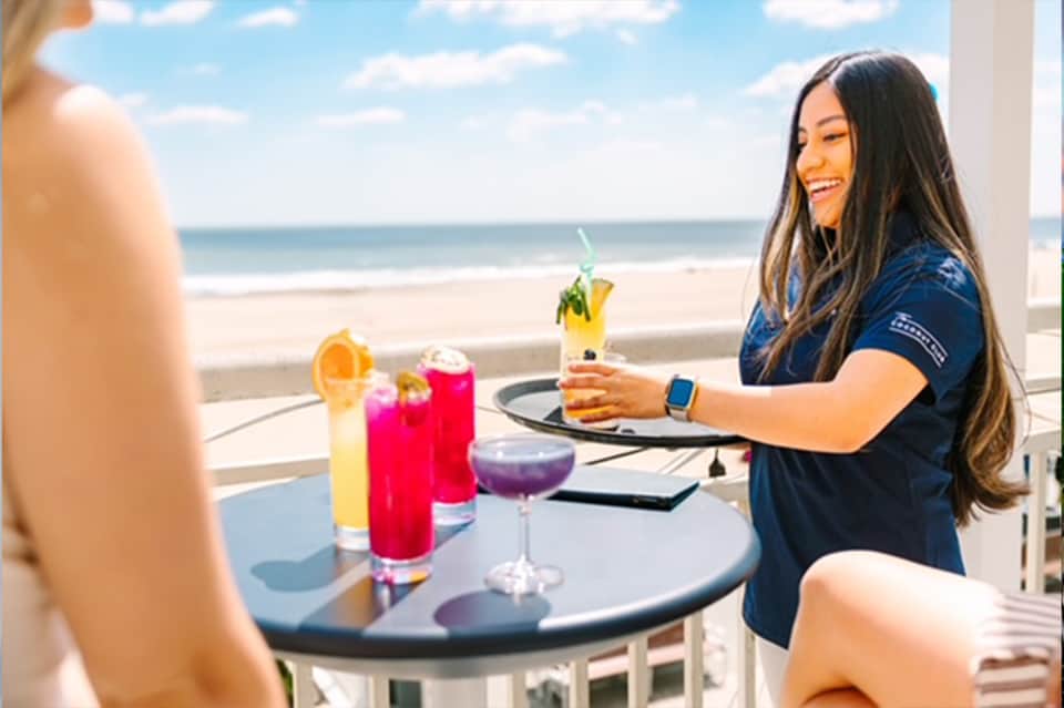 Woman server delivering drinks on beachfront balcony overlooking ocean with colorful cocktails