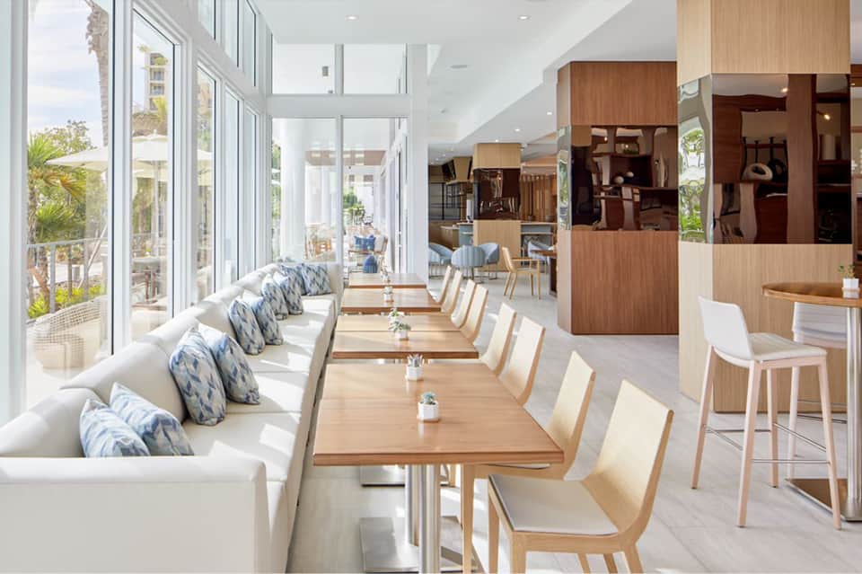 Modern dining room with white banquette seating, wooden tables, and floor-to-ceiling windows overlooking palm trees