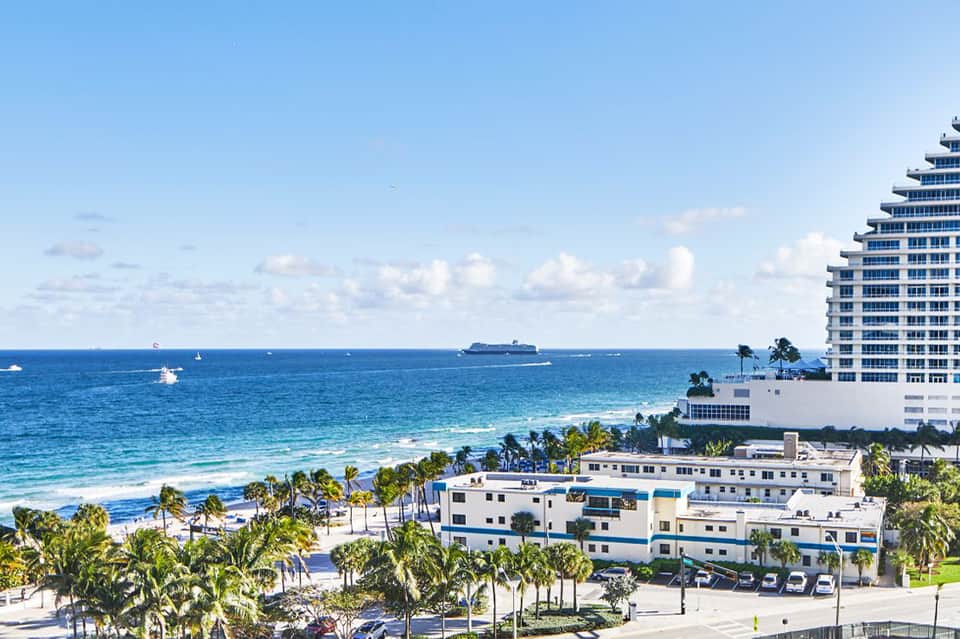 Beachfront resort with high-rise hotel building, palm trees, and cruise ship visible in the ocean under blue sky
