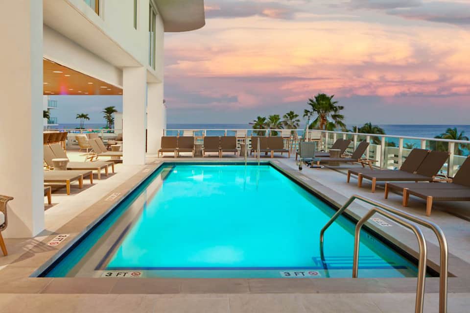 Indoor lap pool with turquoise water, lounge chairs, ocean view, and sunset sky