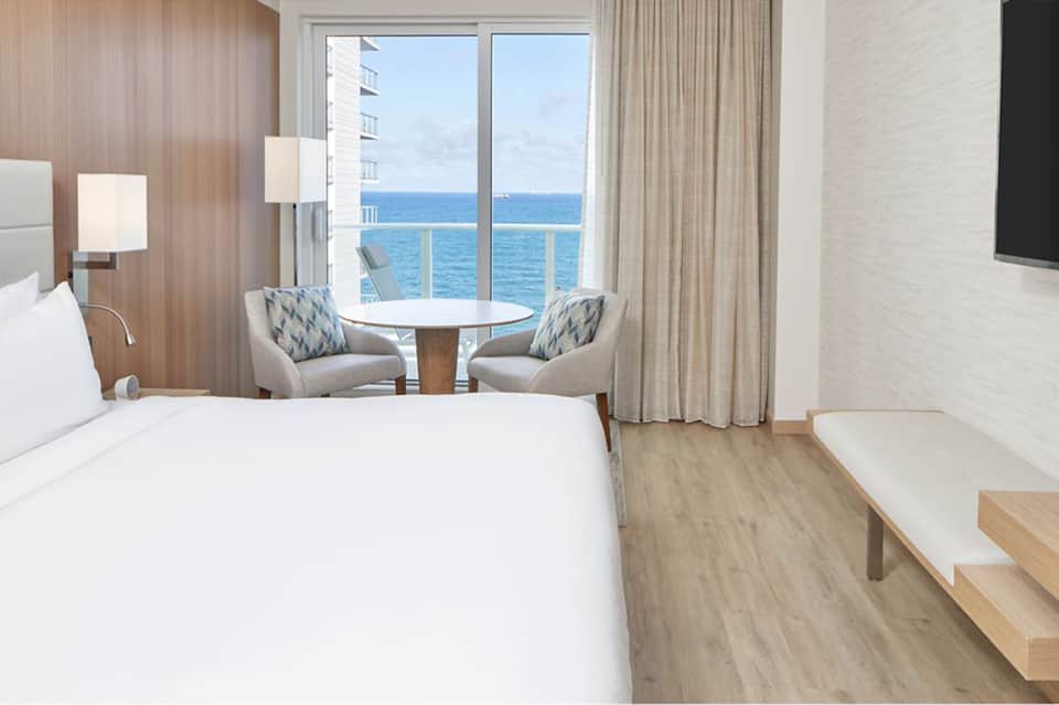 Hotel bedroom with ocean view balcony, modern wood accents, and seating area by windows