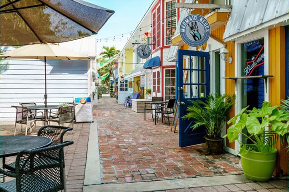 Colorful beachfront shopping district with red, yellow, and blue storefronts, brick pathway, and outdoor seating