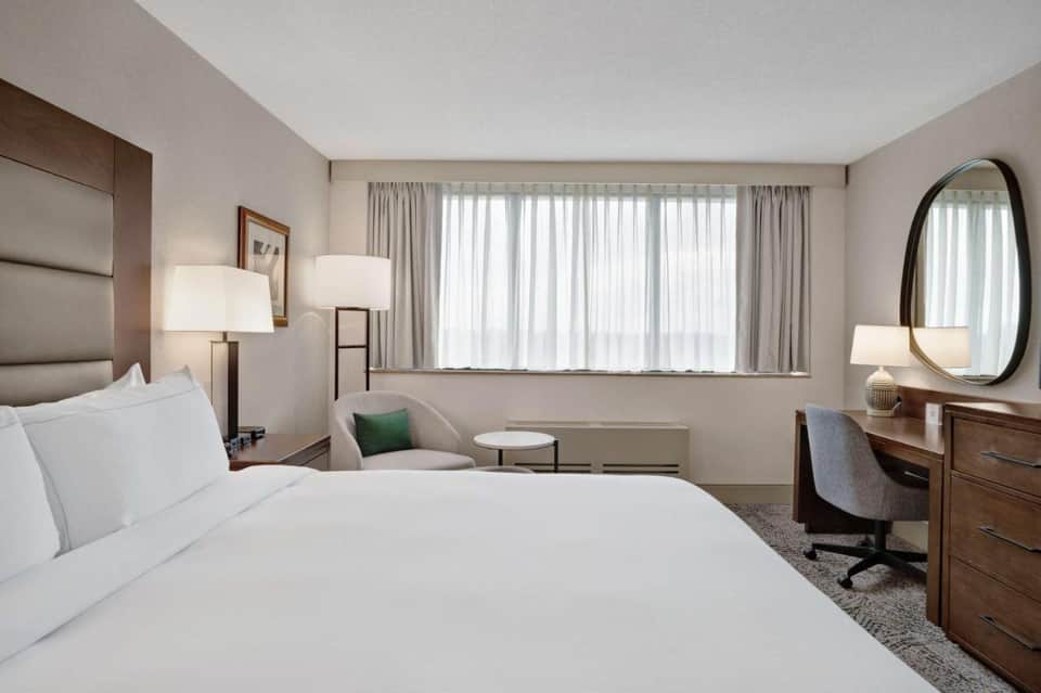 Spacious hotel bedroom with queen bed, work desk, armchair, and large windows with sheer curtains