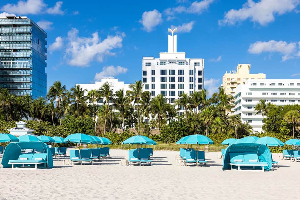 Beach cabanas with turquoise umbrellas and loungers facing Art Deco buildings and palm trees under blue sky