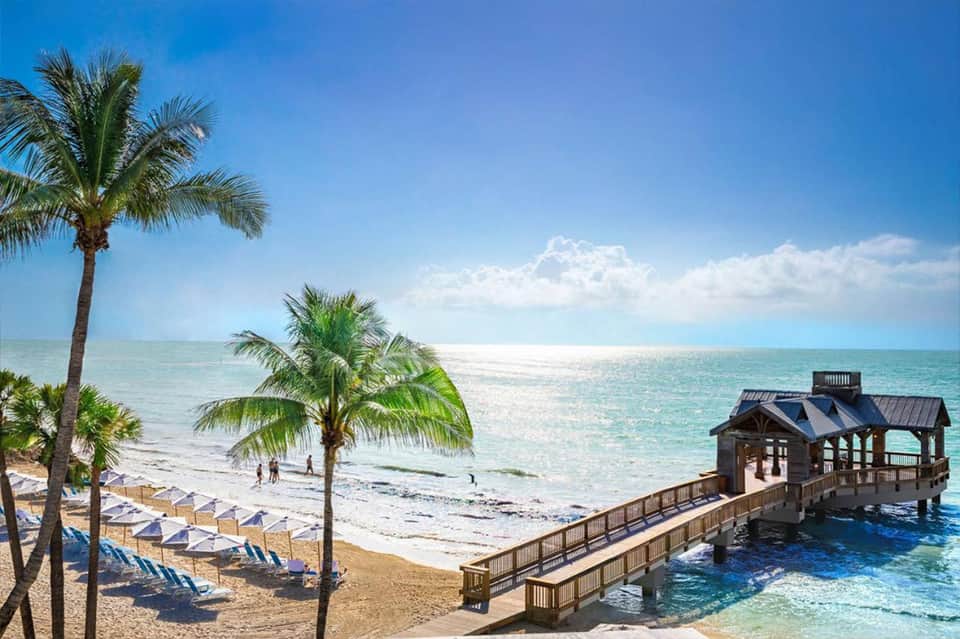 Tropical beach resort with palm trees, sandy shore, lounge chairs, and wooden pier extending into turquoise water