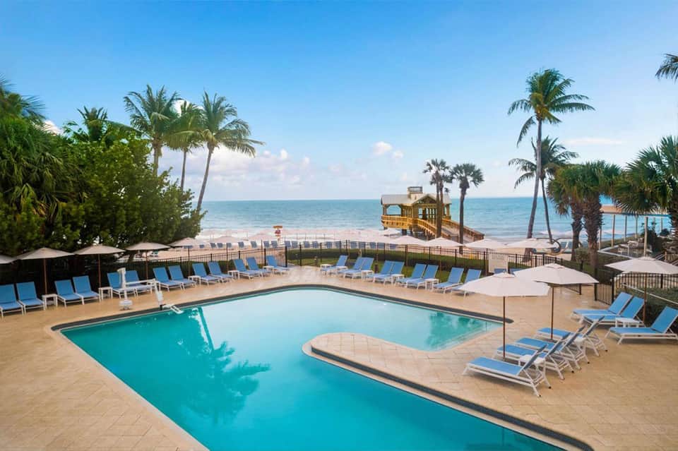 Beachfront resort pool with blue loungers, palm trees, ocean view, and wooden pier