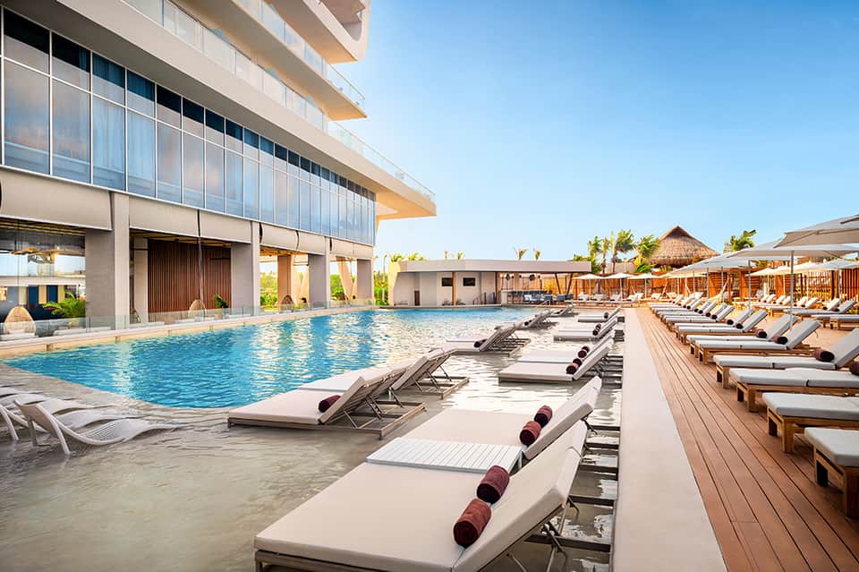 Resort pool deck with rows of cushioned lounge chairs, indoor swimming pool, and modern building architecture