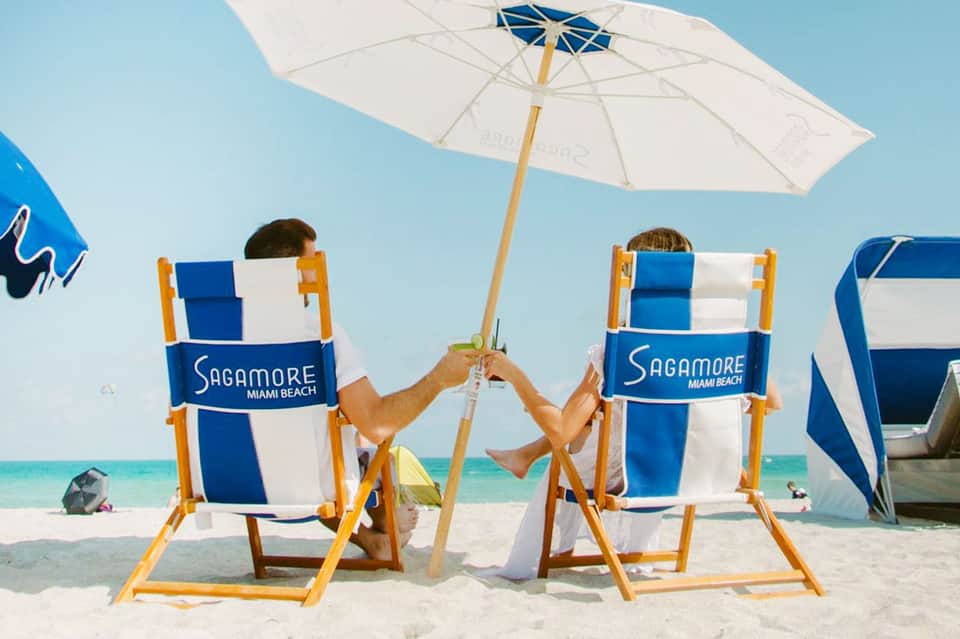 Two people relaxing in Sagamore Miami Beach branded lounge chairs under a white umbrella on sandy beach