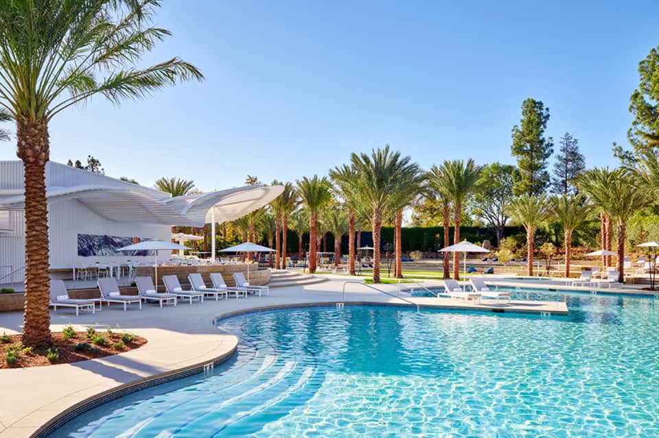 Resort pool area with palm trees, lounge chairs, umbrellas, and modern pavilion under clear sky