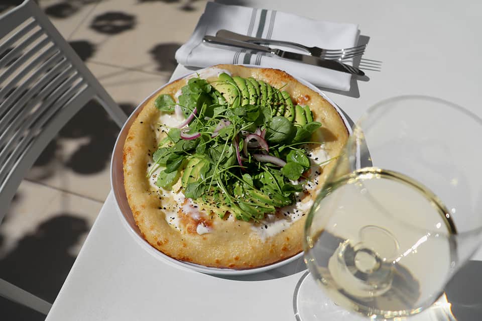 Flatbread pizza topped with greens and vegetables, served with white wine on sunny terrace