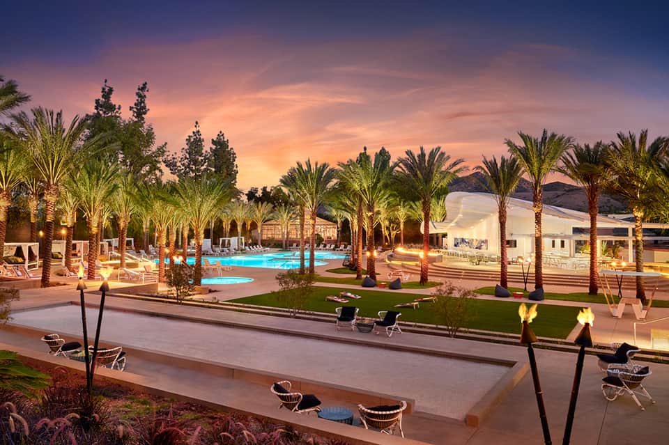 Resort pool at dusk with palm trees, lounge chairs, and modern buildings lit by evening light