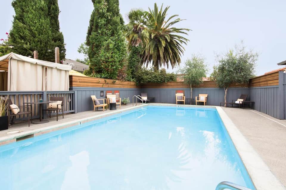 Resort swimming pool with wooden deck enclosed by gray fence, surrounded by lounge chairs and palm trees