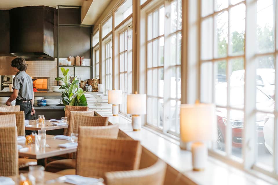 Contemporary restaurant interior with floor-to-ceiling windows, wicker chairs, and open kitchen