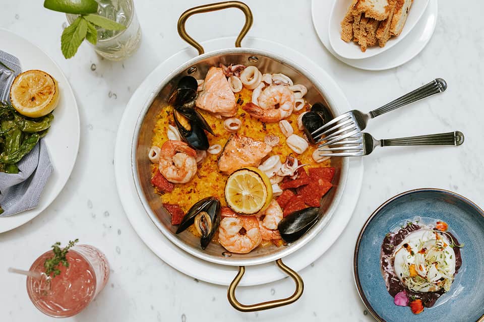 Seafood paella in brass pan with shrimp, mussels, and lemon garnish, surrounded by appetizers