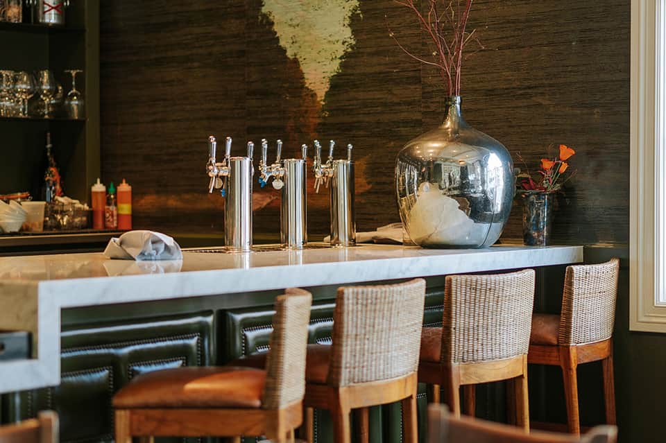 Upscale bar with dark wood accent wall, white countertop, chrome beer taps, and woven dining chairs with leather seats