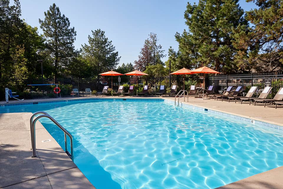Outdoor resort pool with clear blue water, lounge chairs, orange umbrellas, and pine trees