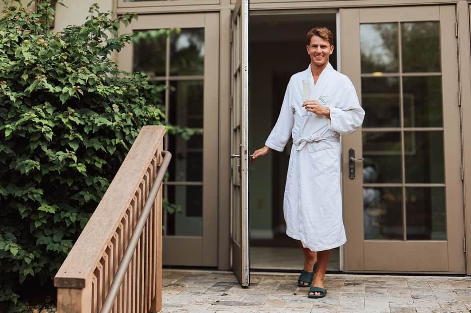 Man in white spa robe standing on outdoor patio with glass doors and lush green ivy-covered walls