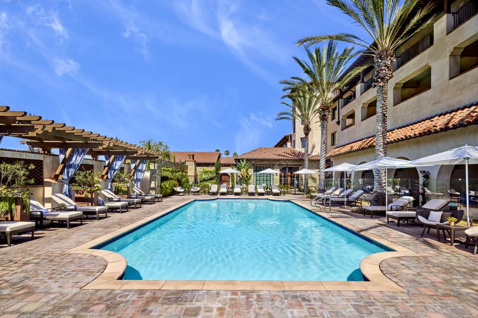 Rectangular lap pool surrounded by lounge chairs, pergolas, and Spanish colonial architecture