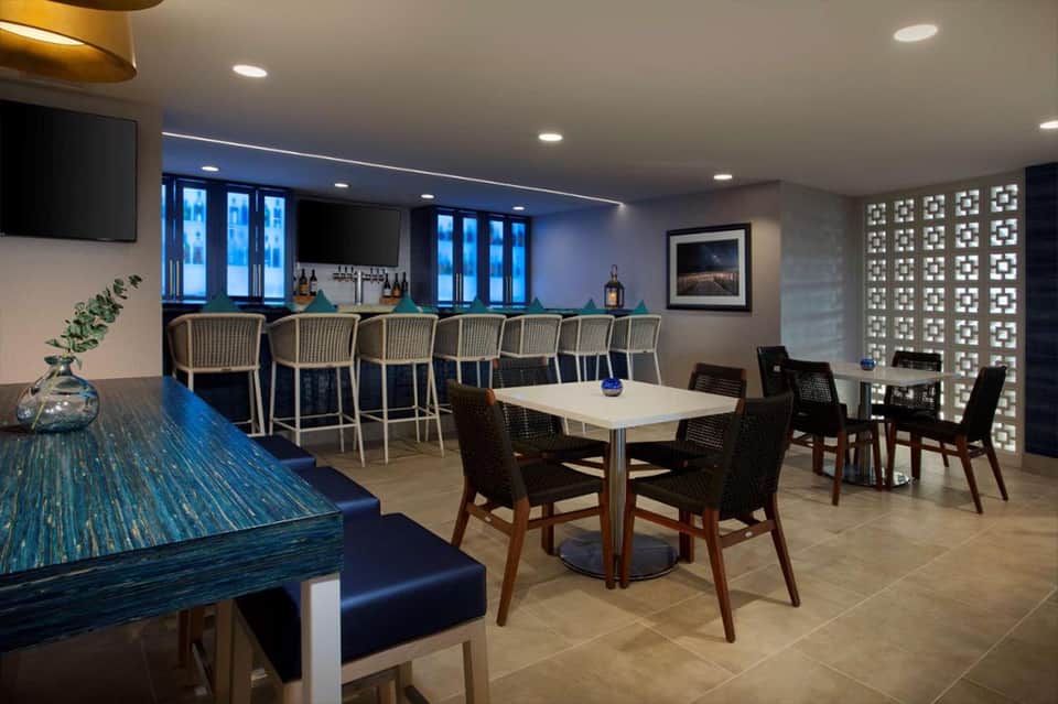 Modern hotel lounge with bar seating, dining tables, teal accent wall, and contemporary furnishings