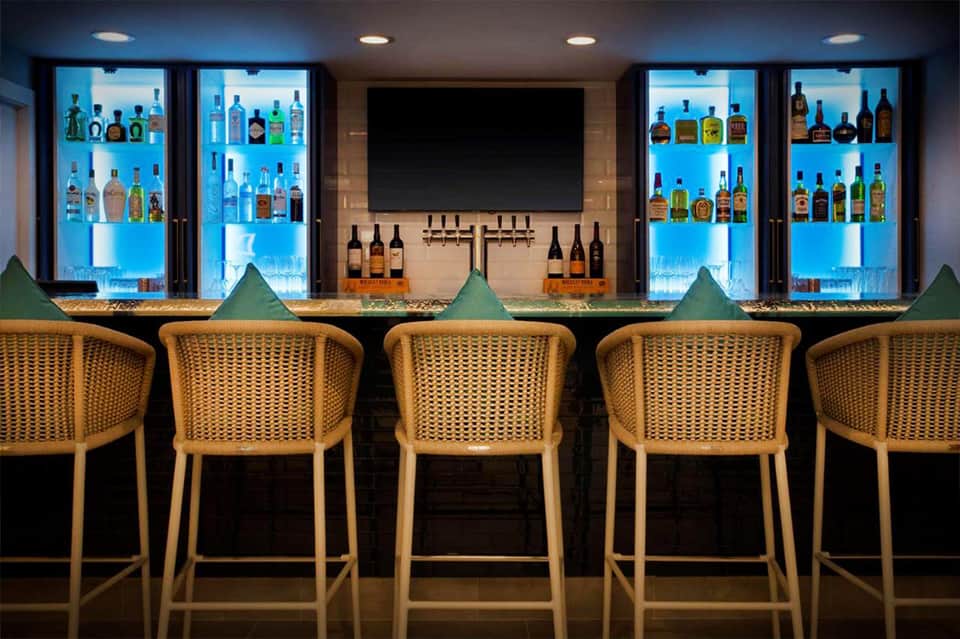 Modern bar with illuminated blue shelving displaying liquor bottles and golden woven bar stools at counter