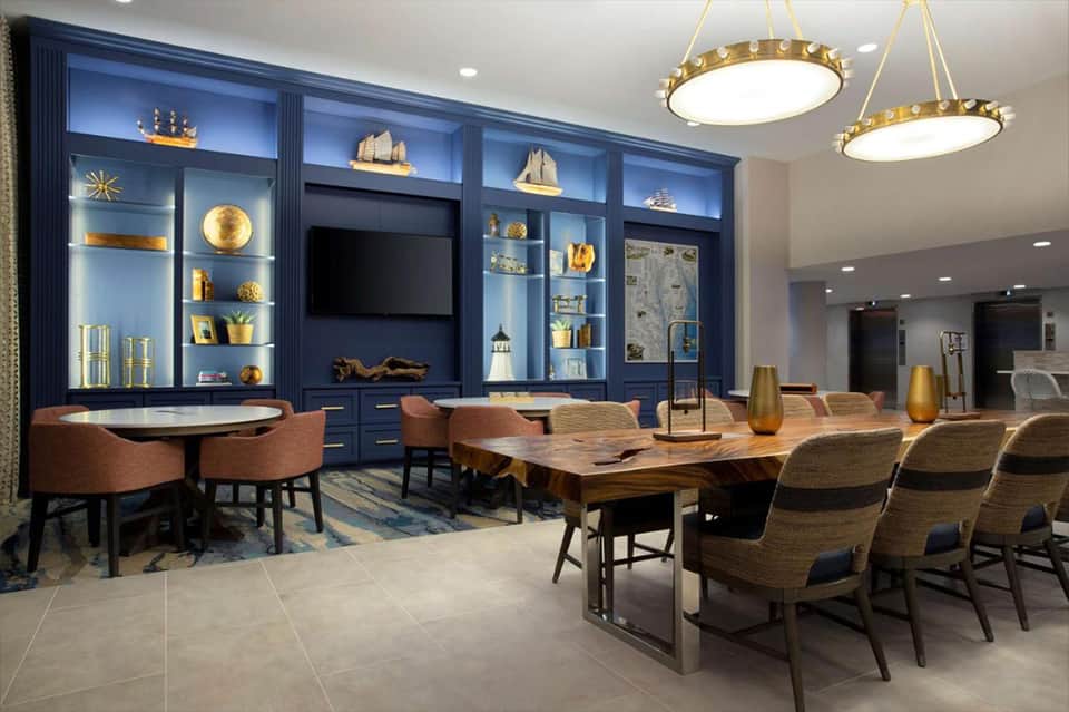 Elegant lounge with blue cabinetry, nautical décor, gold accents, wooden dining table, and brass pendant lights