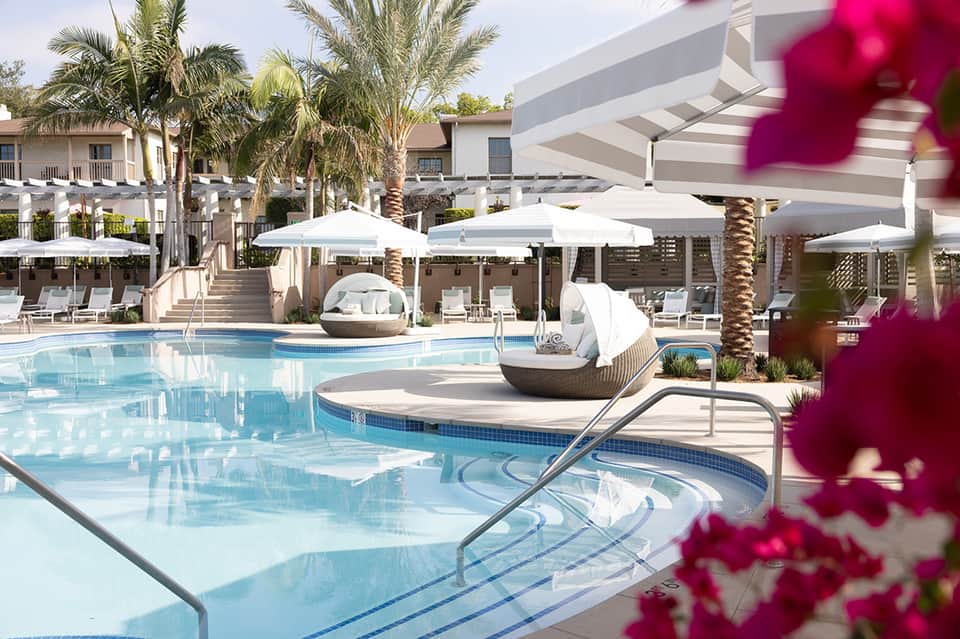 Resort pool area with white umbrellas, palm trees, lounge chairs, and modern white architecture