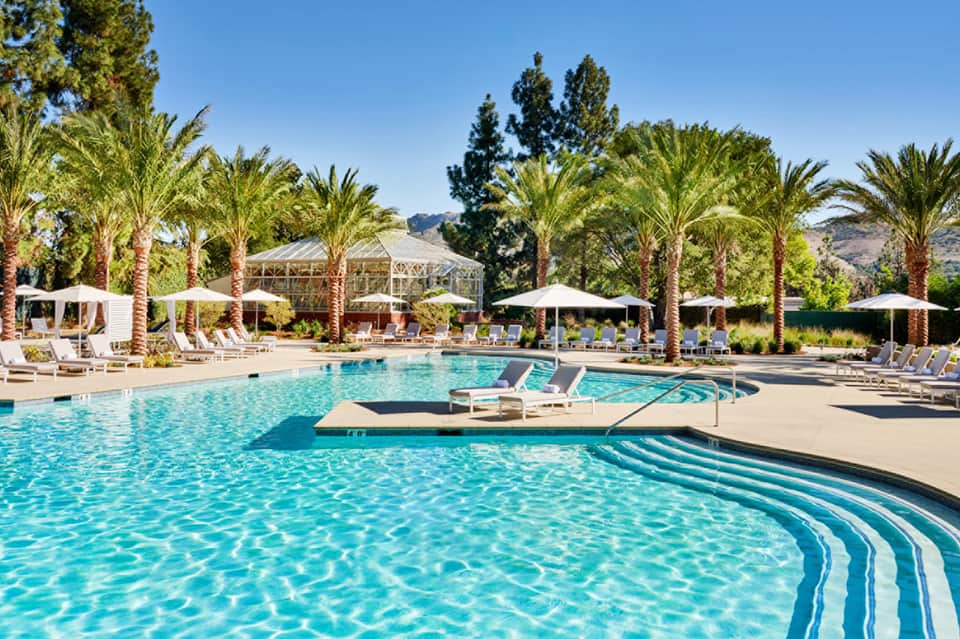 Resort pool with palm trees, lounge chairs, white umbrellas, and mountain views under clear sky