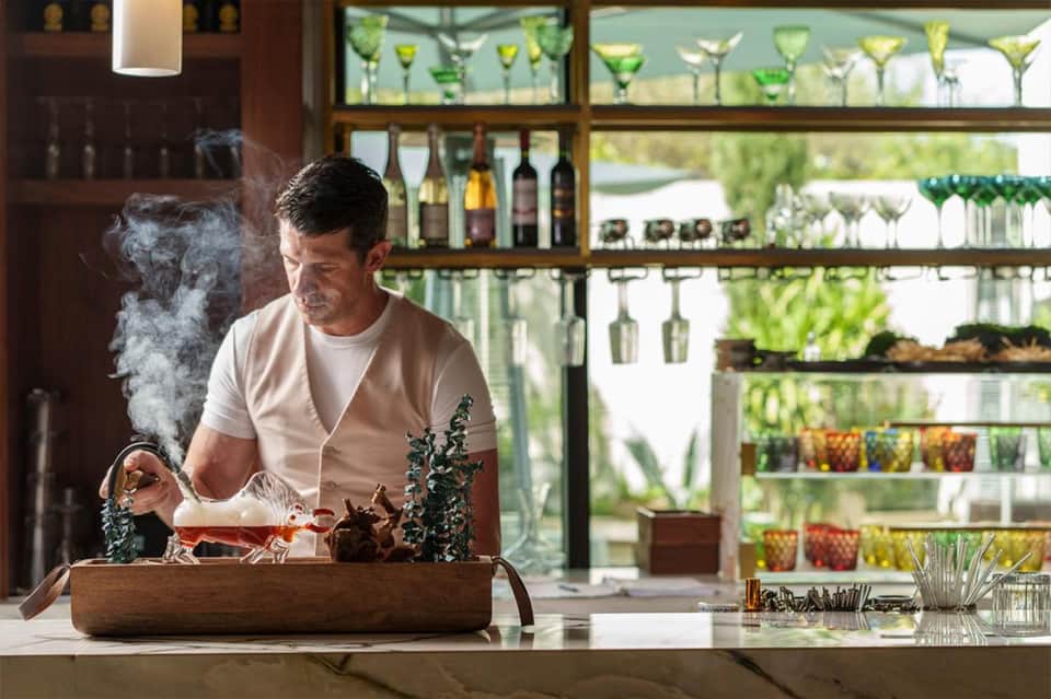 Man performing tea ceremony at luxury bar with botanical garden views and backlit glassware shelving