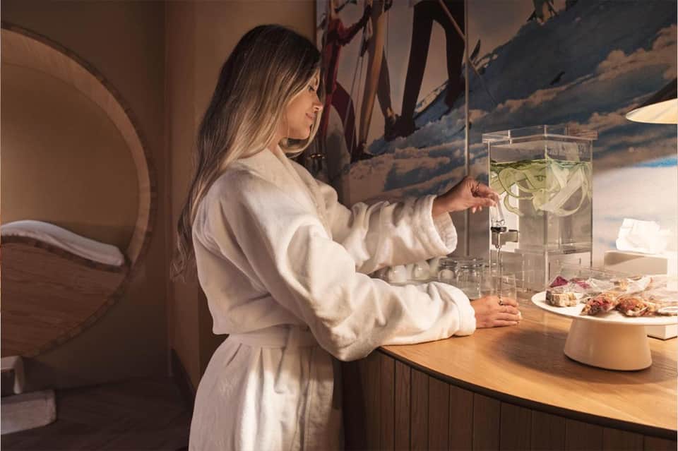 Woman reaching for infused water dispenser at modern wellness bar with minimalist design and snacks