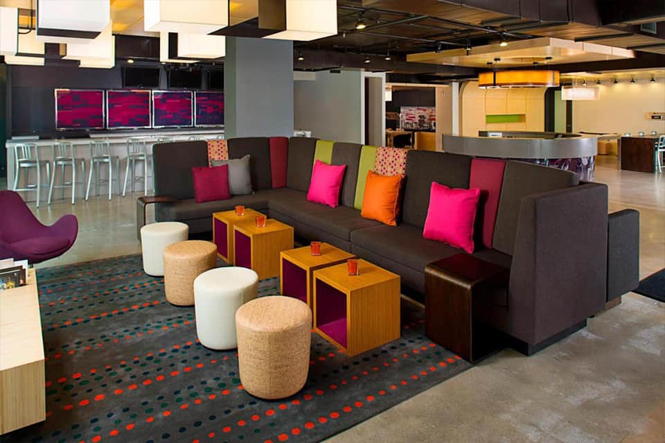 Modern lobby lounge with dark sofa, colorful pillows, cork ottomans, and patterned carpet with bar area
