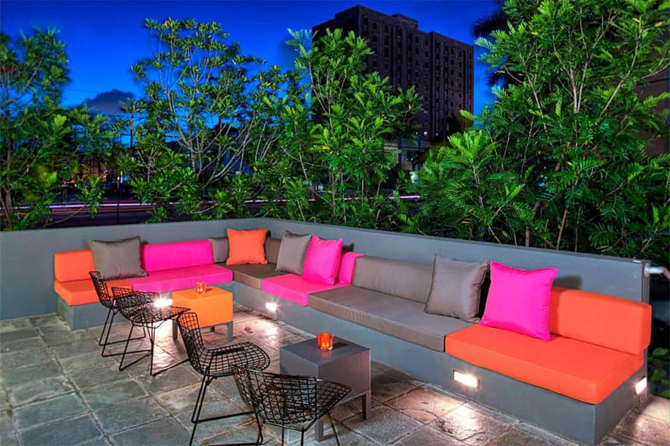 Rooftop lounge at dusk with colorful cushions, potted plants, and city skyline