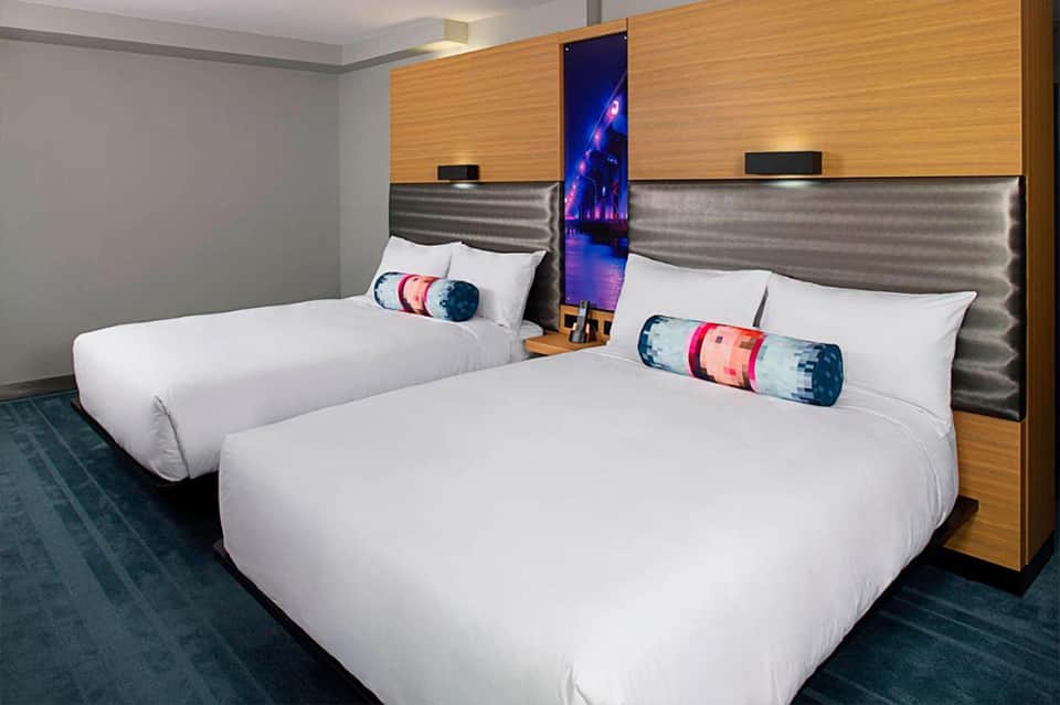 Hotel room with two queen beds, wood headboards, metallic accents, and decorative throw pillows