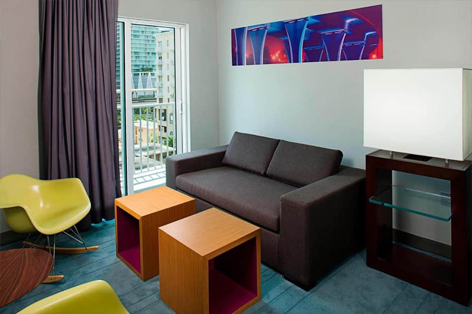 Contemporary hotel suite with dark sofa, yellow accent chair, wooden side tables, purple curtains, and balcony access