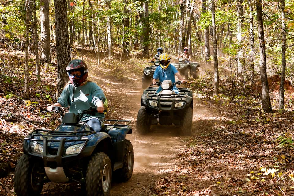 Group of people riding ATVs on a dusty forest trail surrounded by trees