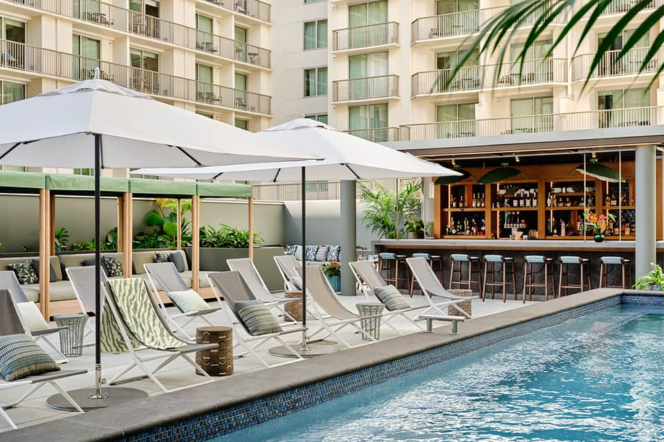 Resort poolside bar with white umbrellas, lounge chairs, wooden bar structure, and palm trees.