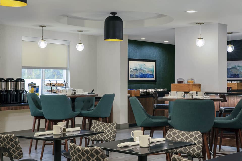 Modern hotel breakfast dining area with teal chairs, pendant lights, and green accent wall featuring artwork