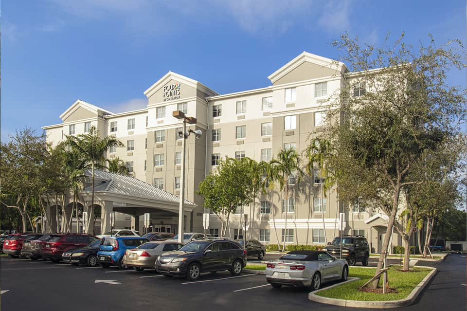 Exterior of beige multi-story hotel with covered entrance, parking lot, palm trees, and landscaping